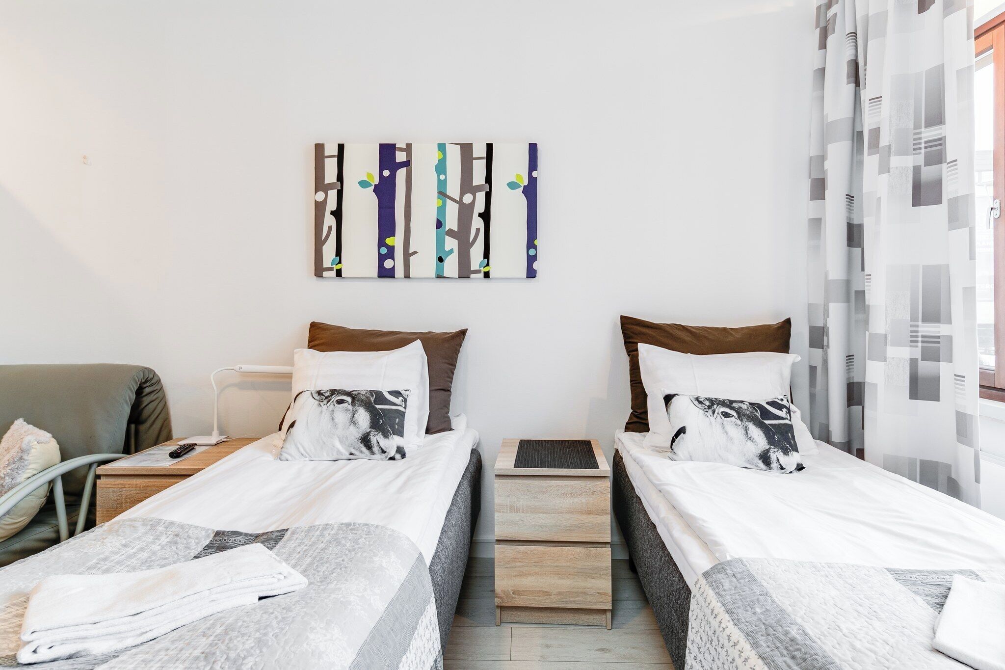 A hotel room with two single beds, a bedside table with a lamp, a chair, and a wall-mounted artwork.