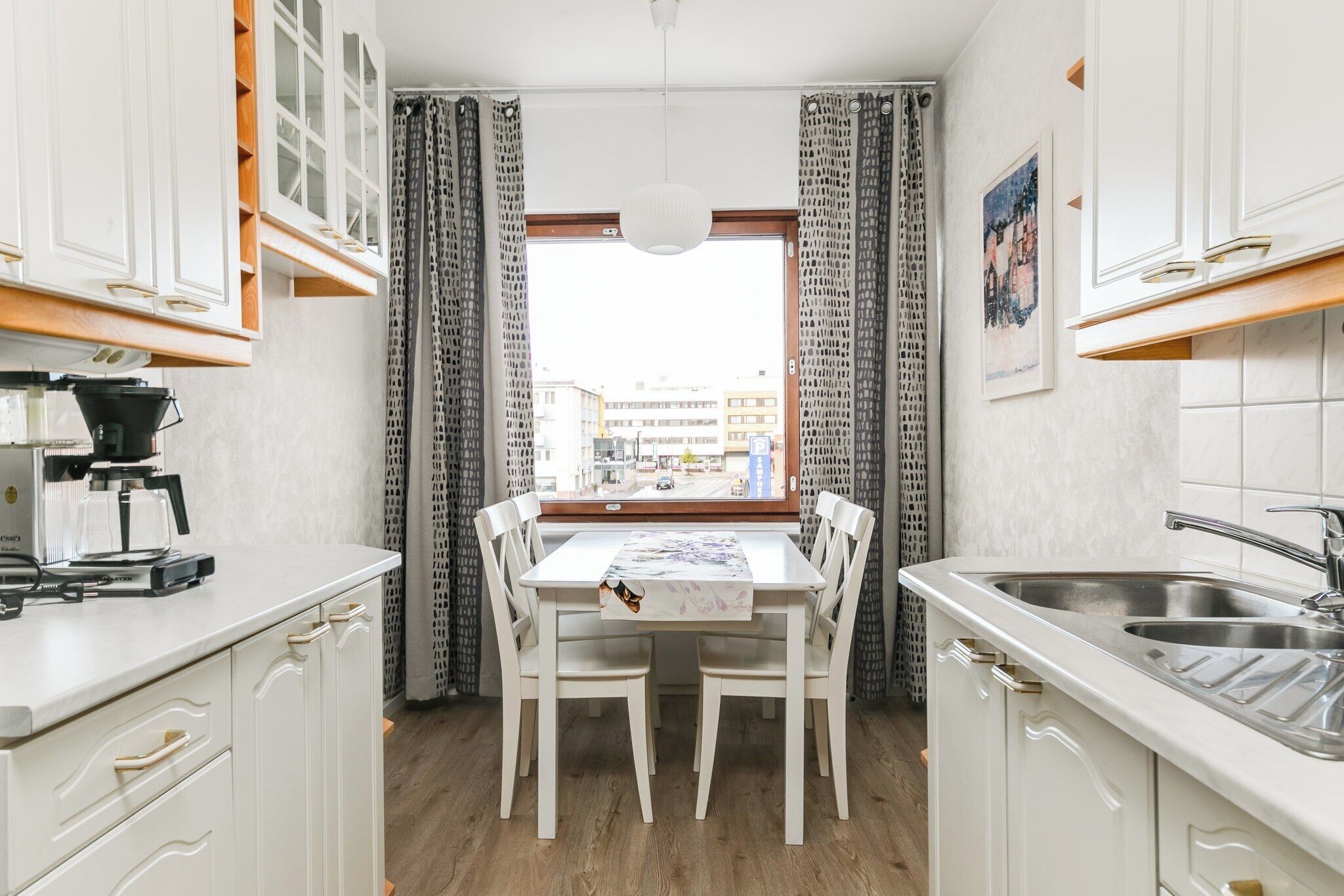 Een kleine keuken met witte kastjes, een eettafel met stoelen, een koffieapparaat en uitzicht door een raam met gordijnen.