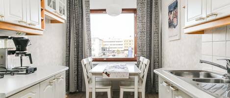 Een kleine keuken met witte kastjes, een eettafel met stoelen, een koffieapparaat en uitzicht door een raam met gordijnen.
