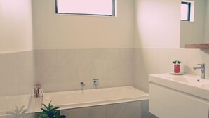 A modern bathroom with a bathtub, a sink, and a mirror. There is a window above the bathtub and a potted plant on the floor. - Relax In True New Modern Style (Christchurch)