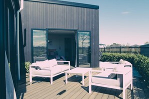 Terraza exterior con mobiliario blanco, incluyendo sofás y una mesa, y un ventanal de vidrio que da a un espacio interior.