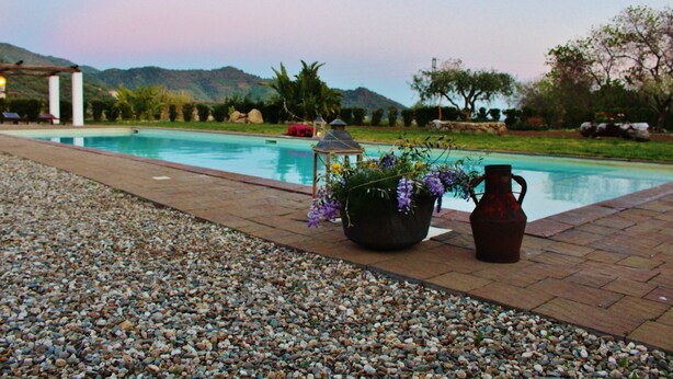 A swimming pool with a pebble border, surrounded by a garden with a gazebo and a large vase with flowers. - Tenuta Edonè Agriturismo (Graniti)