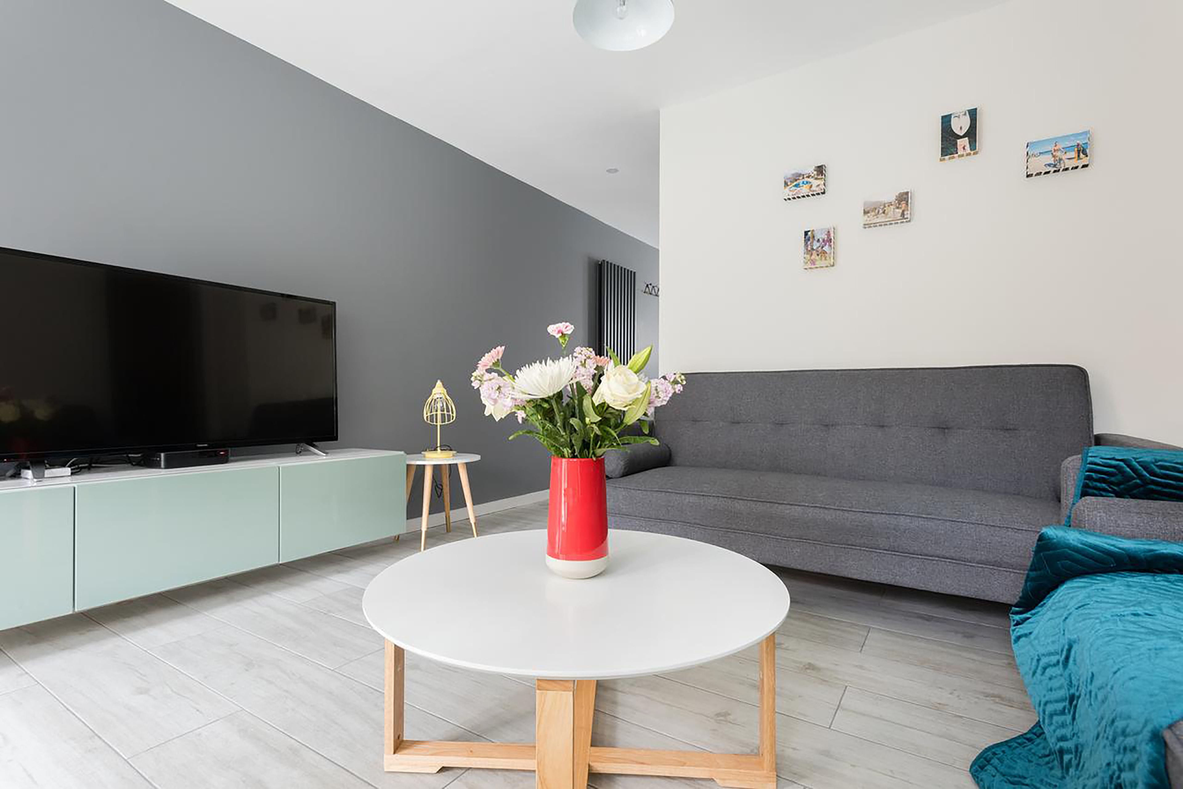 A modern living room with a flat-screen TV, a round white coffee table, a grey sofa, and a red vase with flowers.