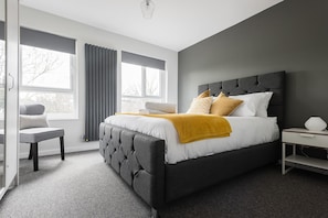 A modern bedroom with a large bed, grey and white bedding, a bedside table, and a chair. - The Waterloo Residence (London)