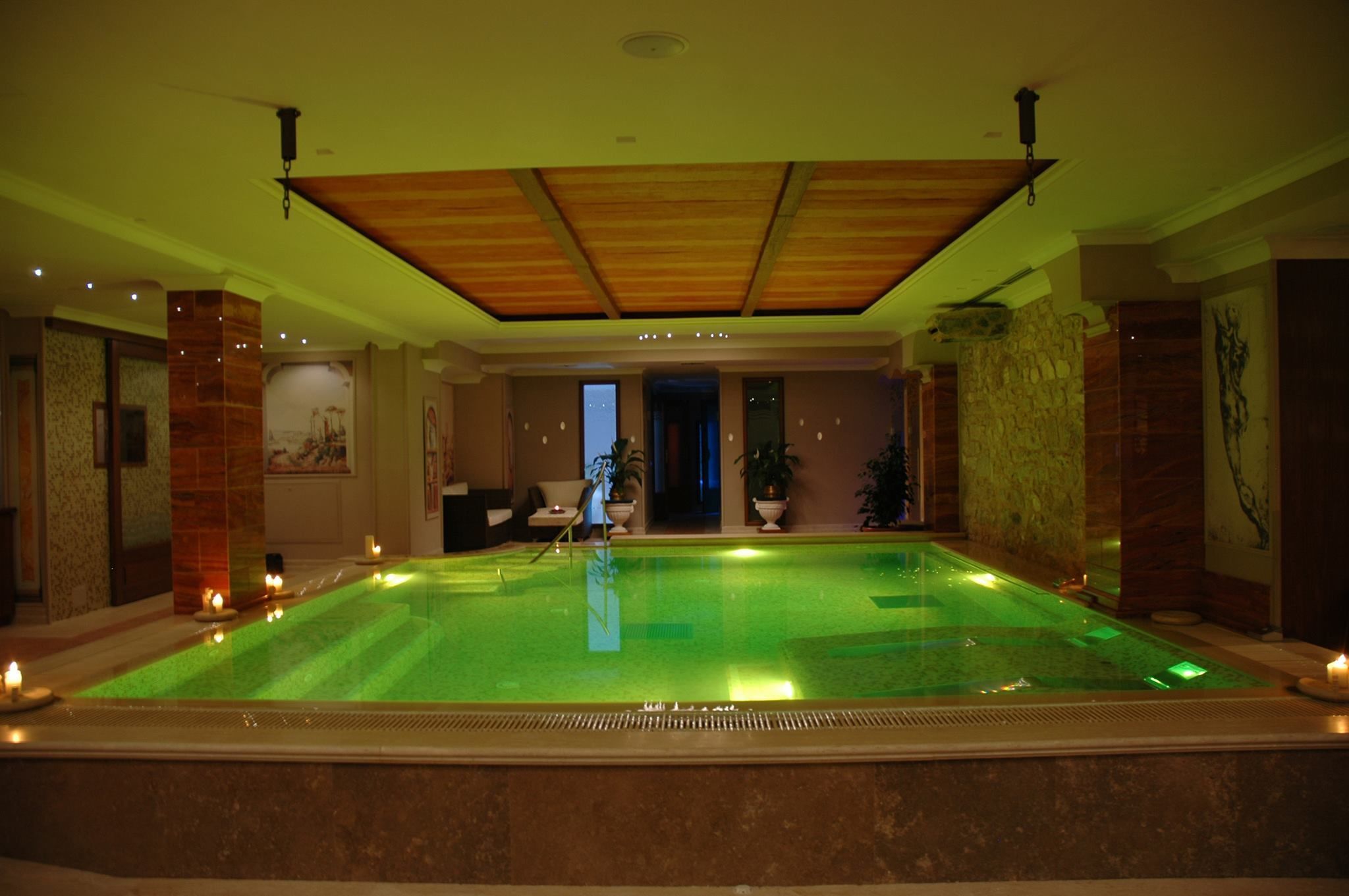 A large indoor swimming pool with a wooden ceiling and stone walls, surrounded by candles and potted plants.