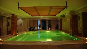 A large indoor swimming pool with a wooden ceiling and stone walls, surrounded by candles and potted plants.