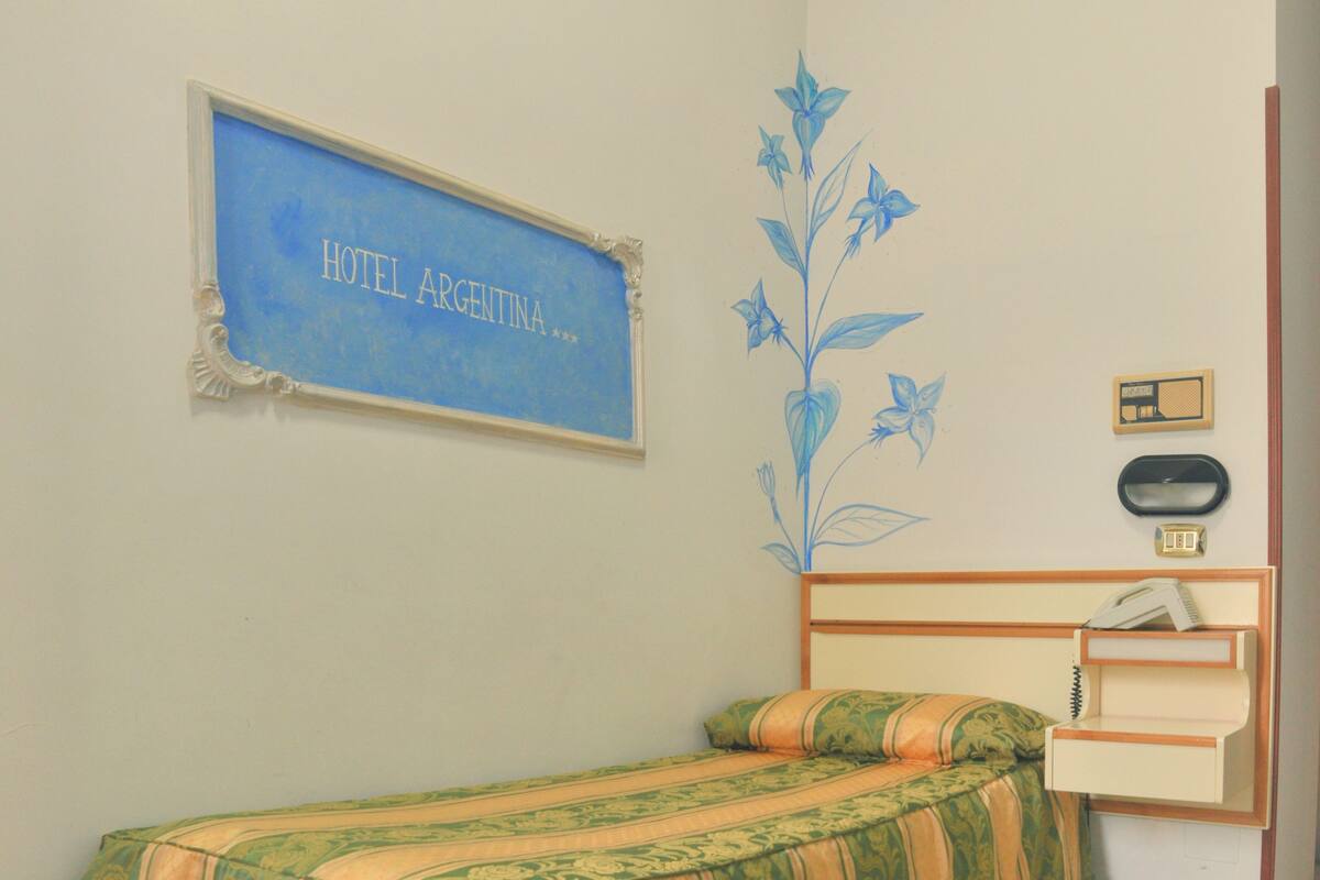 A single bed with a floral bedspread, a small bedside table with a radio, and a framed sign on the wall.
