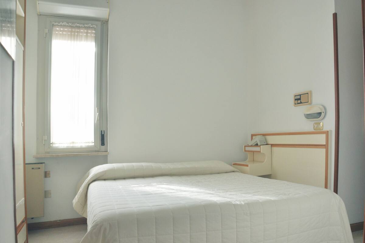 A simple, clean bedroom with a single bed, a bedside table, and a window with curtains.