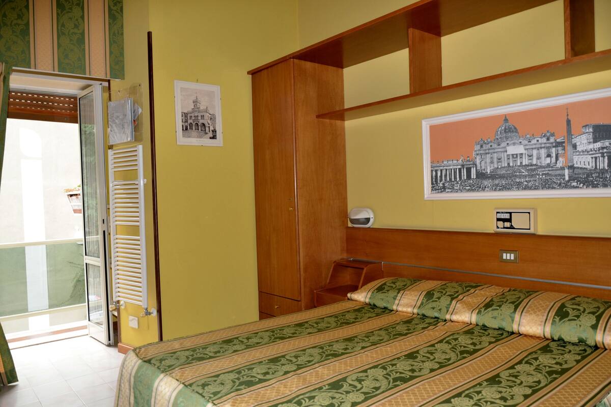 A hotel room with a bed, a wooden wardrobe, a small table, and a wall-mounted shelf.