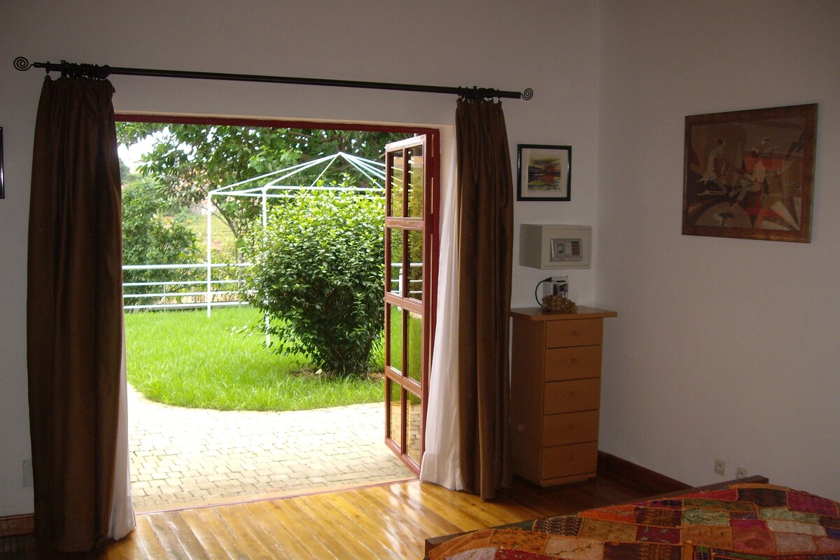 Ein Zimmer mit Holzboden, einem Bett mit bunter Steudecke, einem hölzernen Schminktisch, einem Bild an der Wand und Blick auf einen Garten durch eine geöffnete Tür.