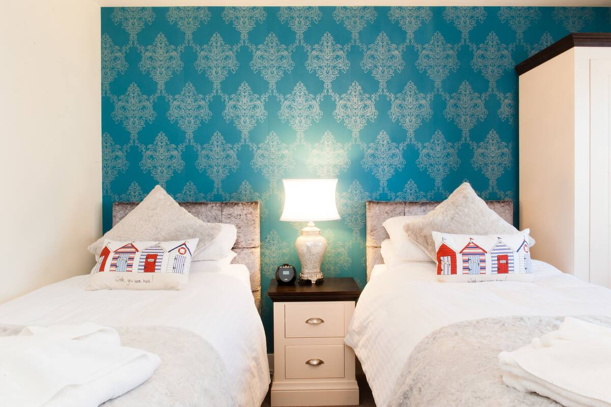 A hotel room with two beds, a nightstand with a lamp, and a patterned wall.