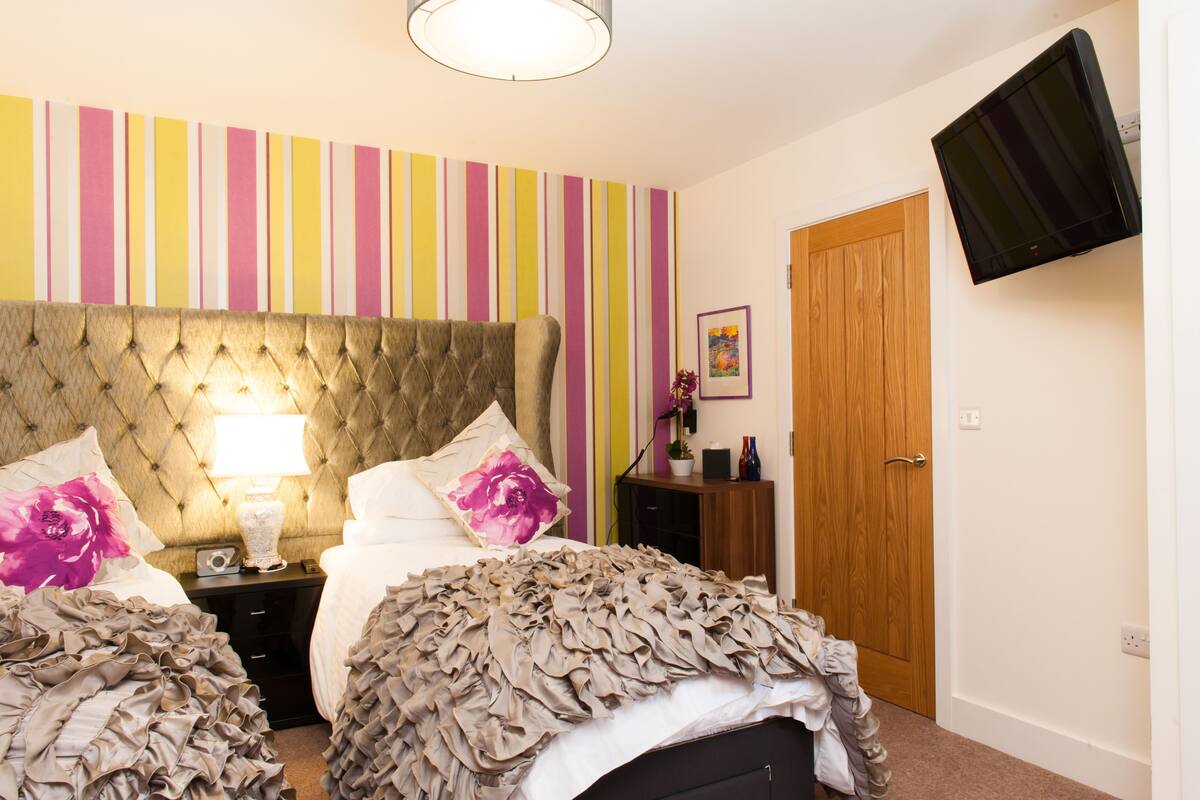 A bedroom with a tufted headboard, two beds with ruffled bedding, a bedside table with a lamp, a TV mounted on the wall, and a wooden door.