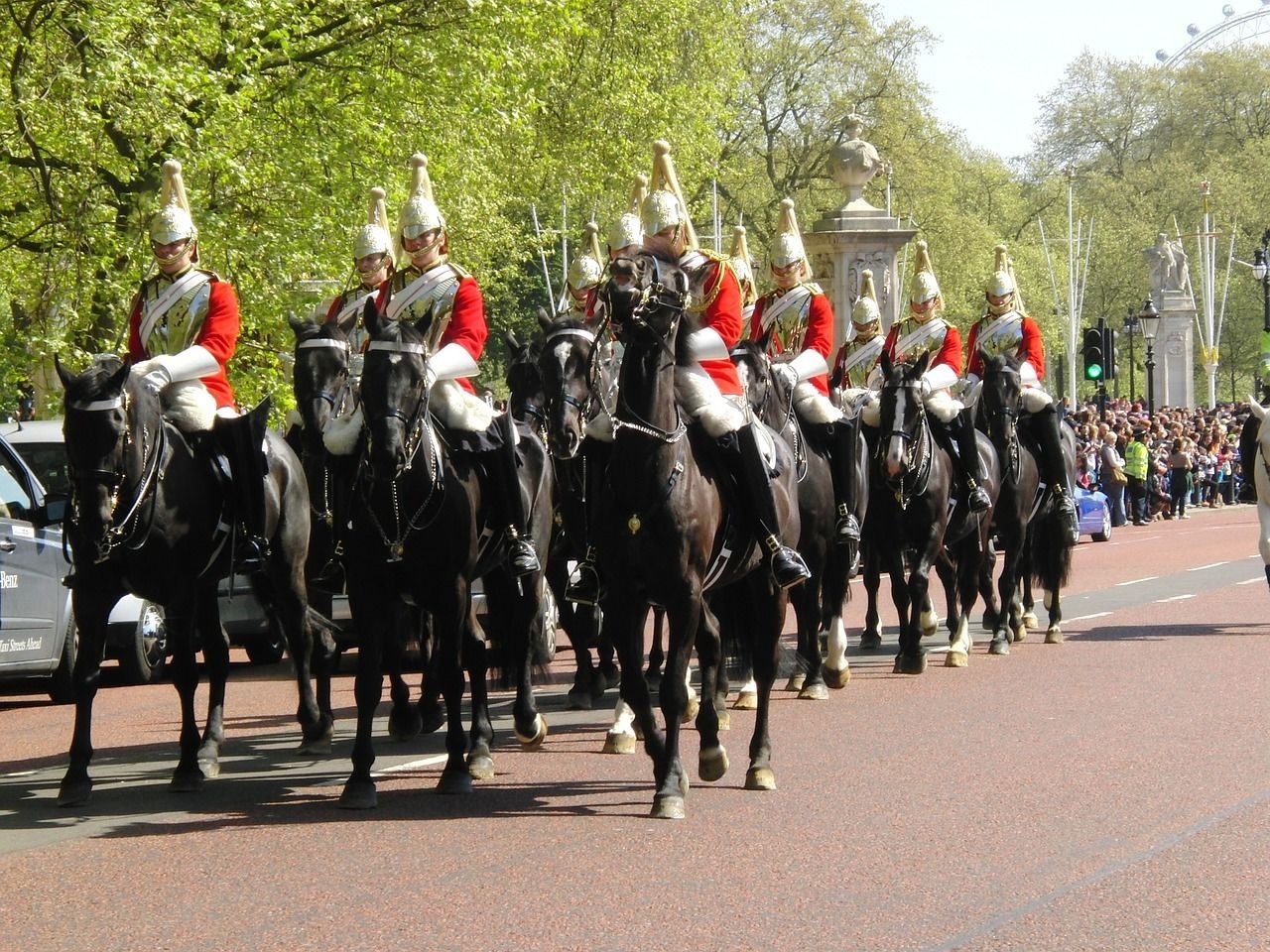 A group of mounted guards in red uniforms with white helmets, walking down a street lined with spectators and trees.