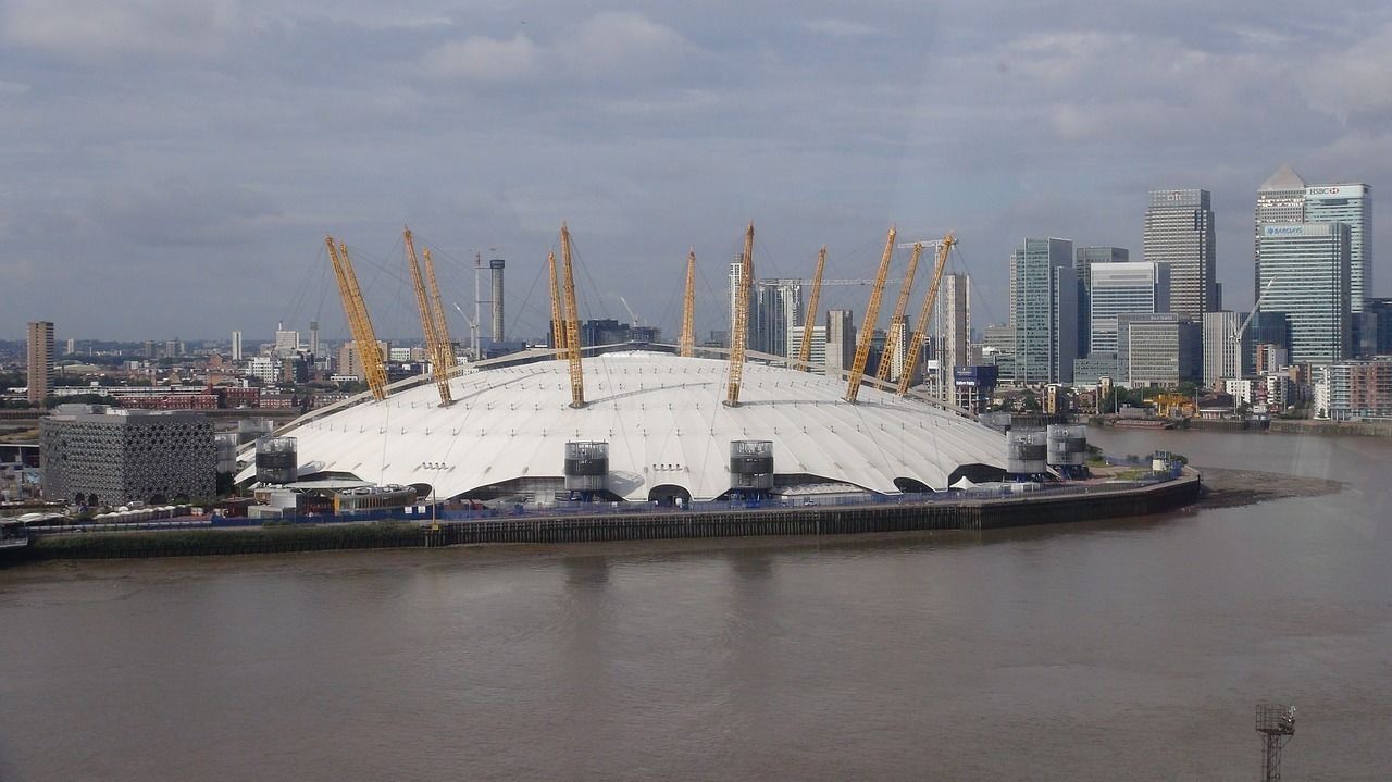 A stadium with a white roof and multiple cranes, surrounded by a city skyline and a body of water.