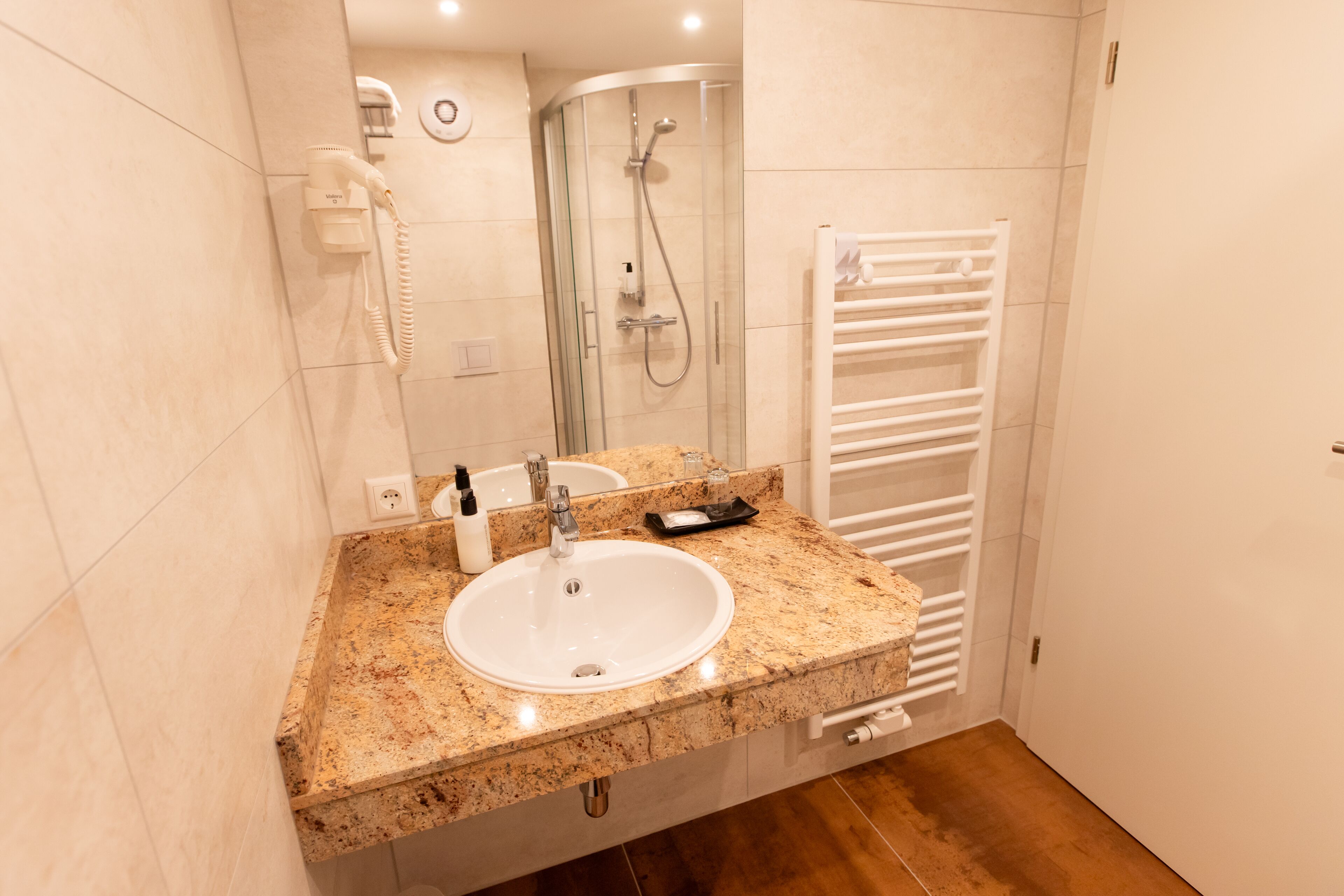 Un baño con lavamanos de encimera de granito, ducha con cerramiento de vidrio y toallero blanco.