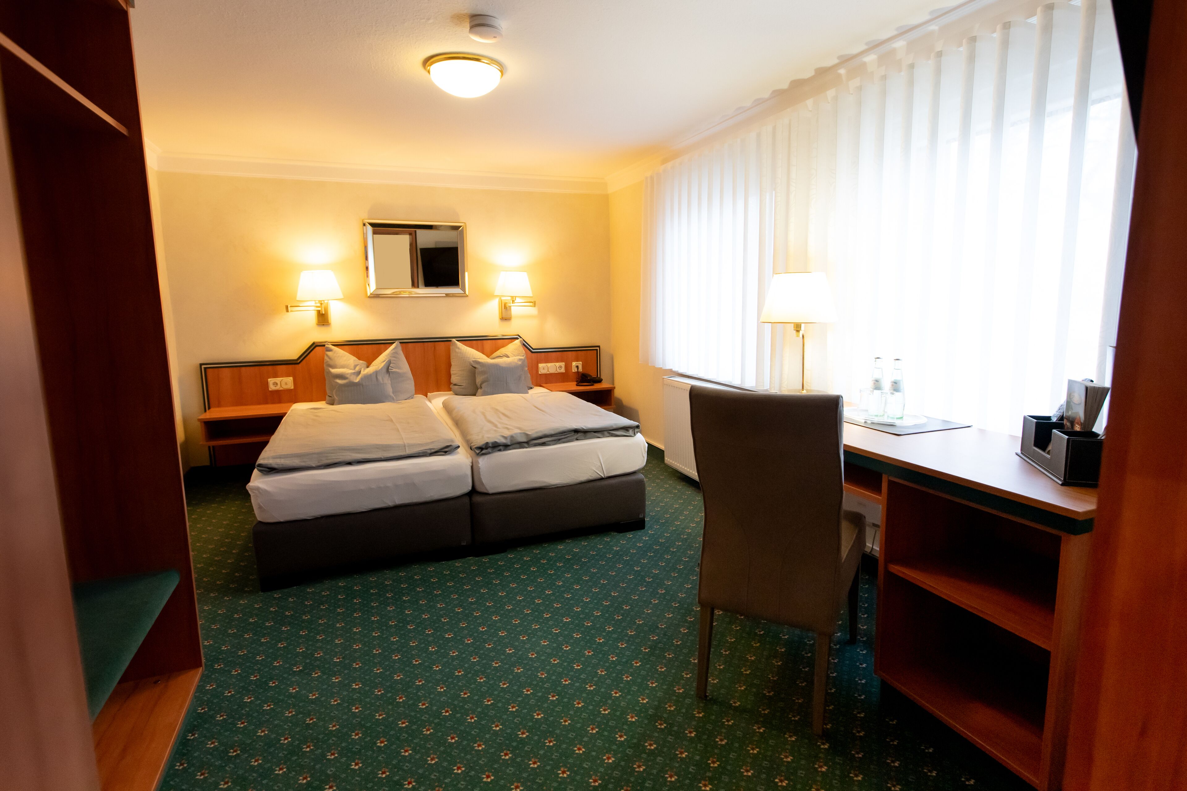 Habitación de hotel con cama doble, dos mesitas de noche, un escritorio, una silla y un amplio ventanal con cortinas.