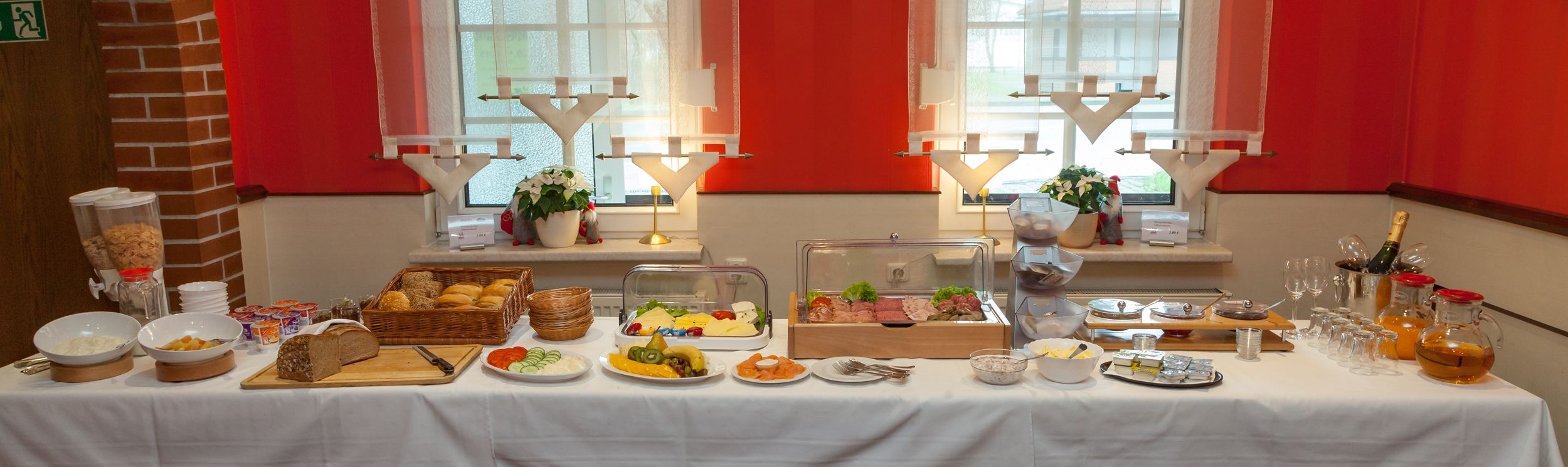 A buffet table with various food items, including bread, fruits, and salads, set against a red wall with windows.