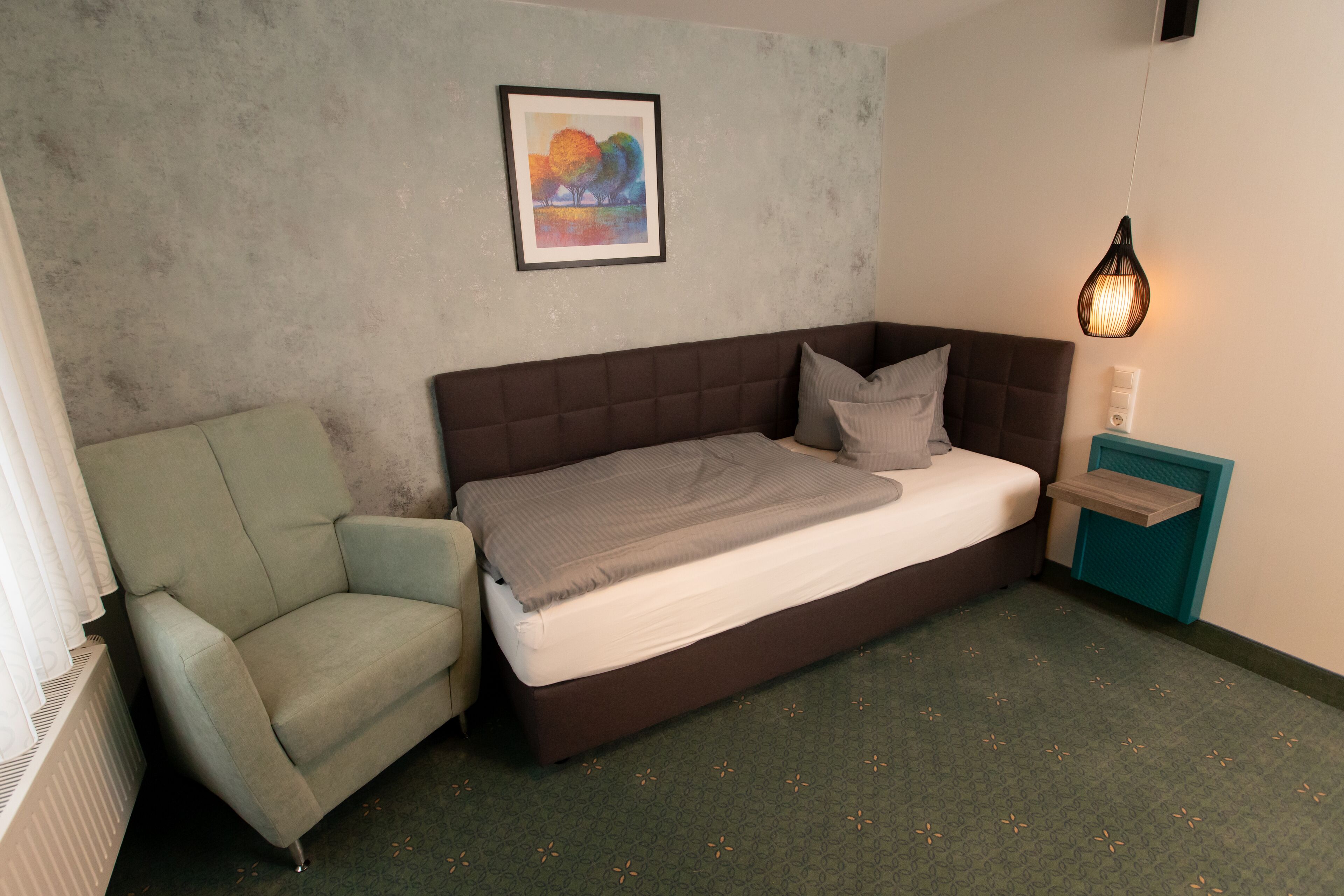 A bedroom with a bed, a chair, a bedside table, a lamp, and a framed picture on the wall.