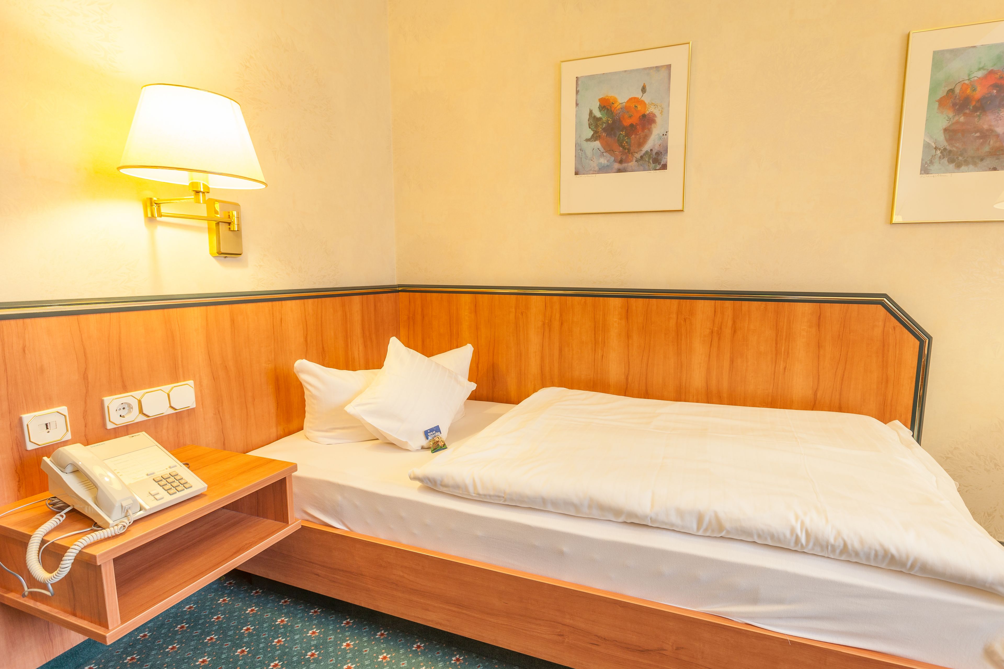 Ein Hotelzimmer mit einem hölzernen Kopfteil, einem Nachttisch mit Telefon, einer Lampe und zwei gerahmten Kunstwerken an der Wand.