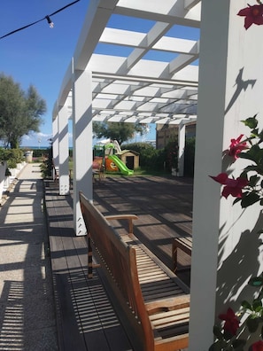 Stroll through the shaded walkway under a picturesque pergola, surrounded by lush greenery and a nearby playground.