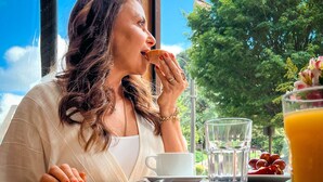 A person sitting at a table with a cup and a glass of orange juice, with a window view of trees and a clear sky. - Hotel Vovó Carolina (Gramado)