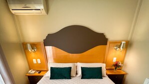 A neatly made bed with a patterned bedspread and two pillows, a wooden headboard, bedside lamps, and an air conditioning unit above the bed. - Hotel Vovó Carolina (Gramado)