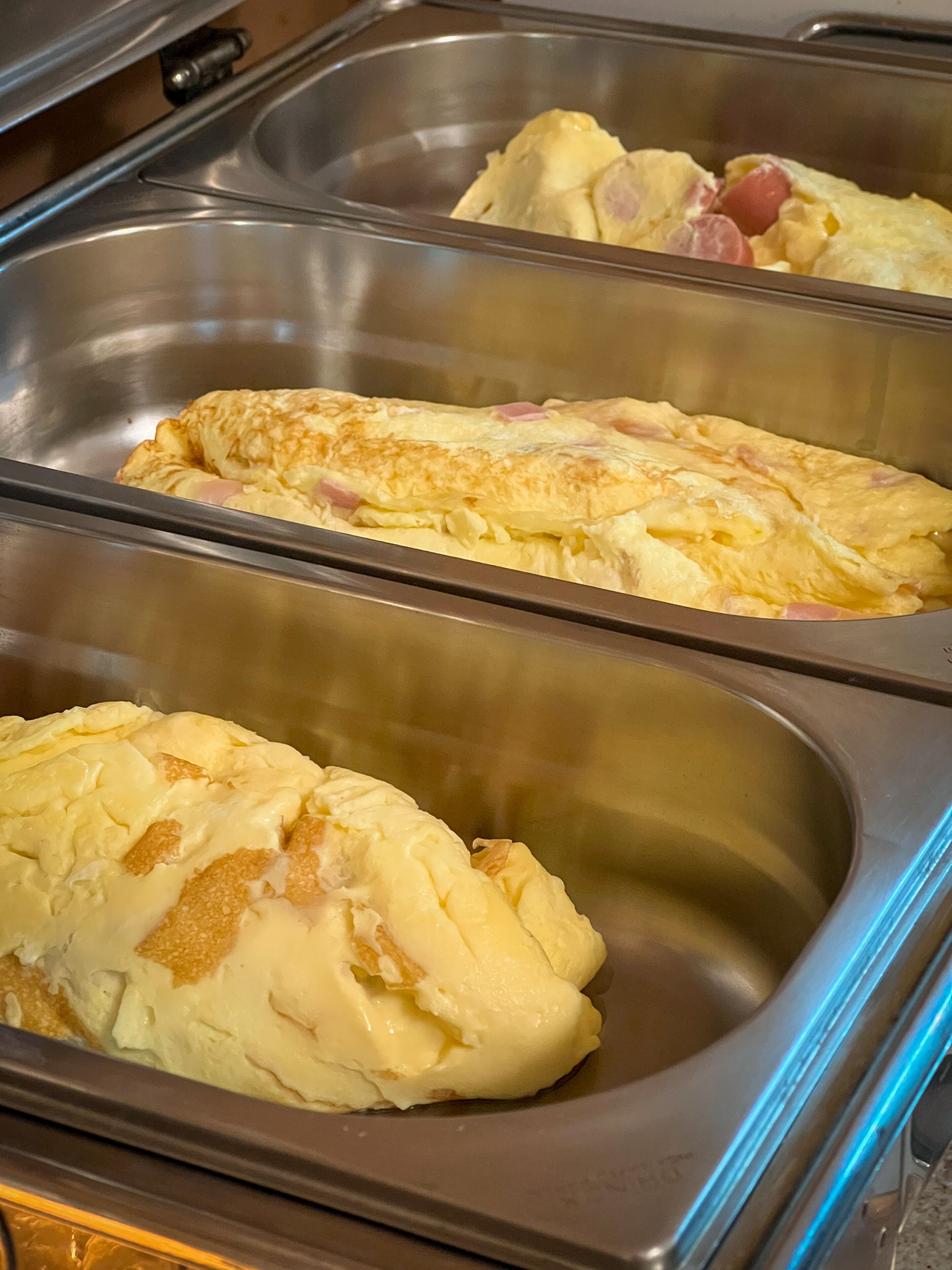 A buffet with omelettes and other dishes in stainless steel serving trays.