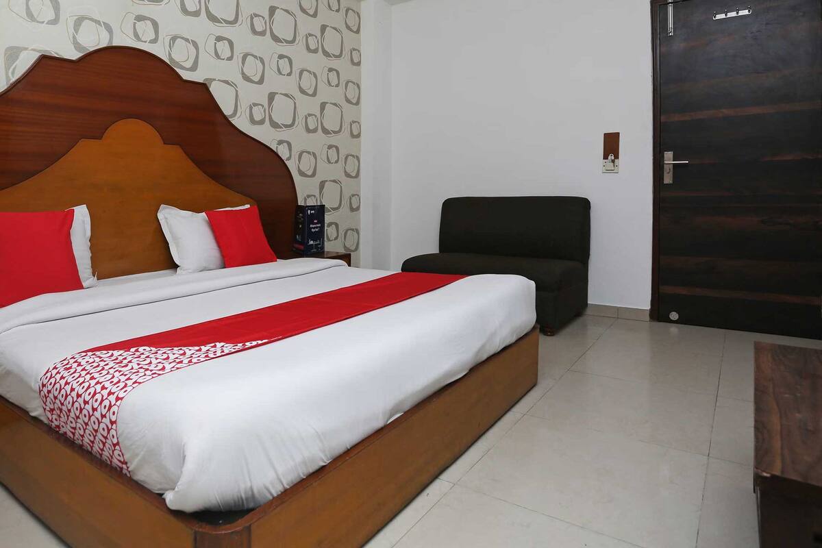 Una cama bien hecha con un cabecero de madera, una mesita de noche con un teléfono y una silla en una habitación con un papel tapiz estampado y una puerta oscura.