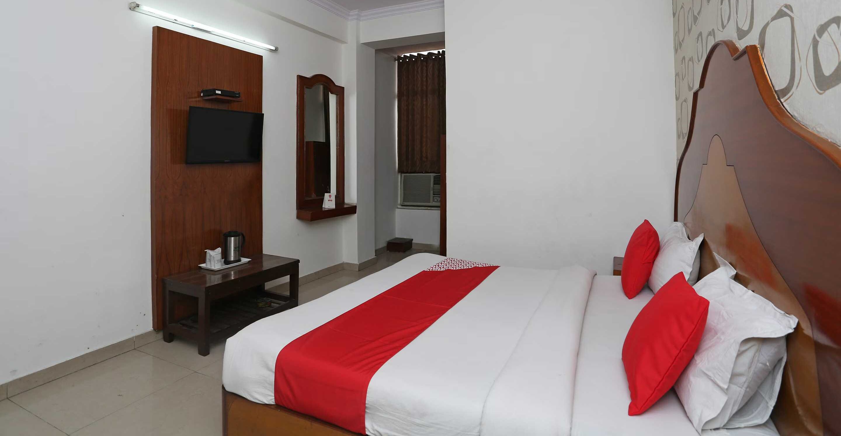 A hotel room with a bed, a TV, a mirror, a small table with a kettle, and a window with curtains.