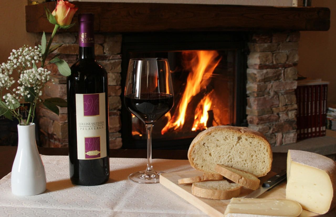 A bottle of wine, a glass of wine, slices of bread, and cheese on a table with a fireplace in the background.