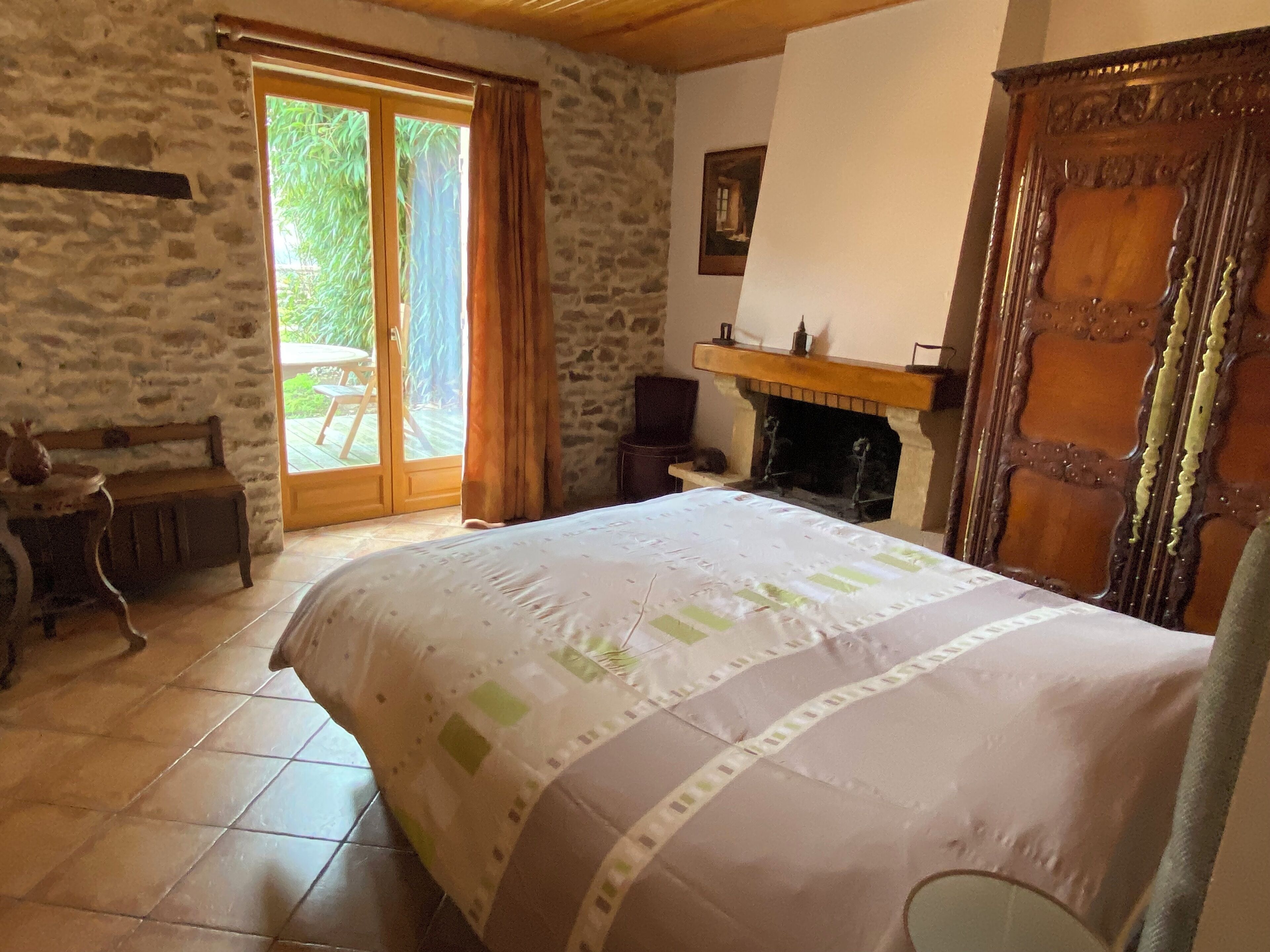 Une chambre avec un mur en pierre, un lit en bois, une cheminée et une porte-fenêtre coulissante donnant sur une terrasse.