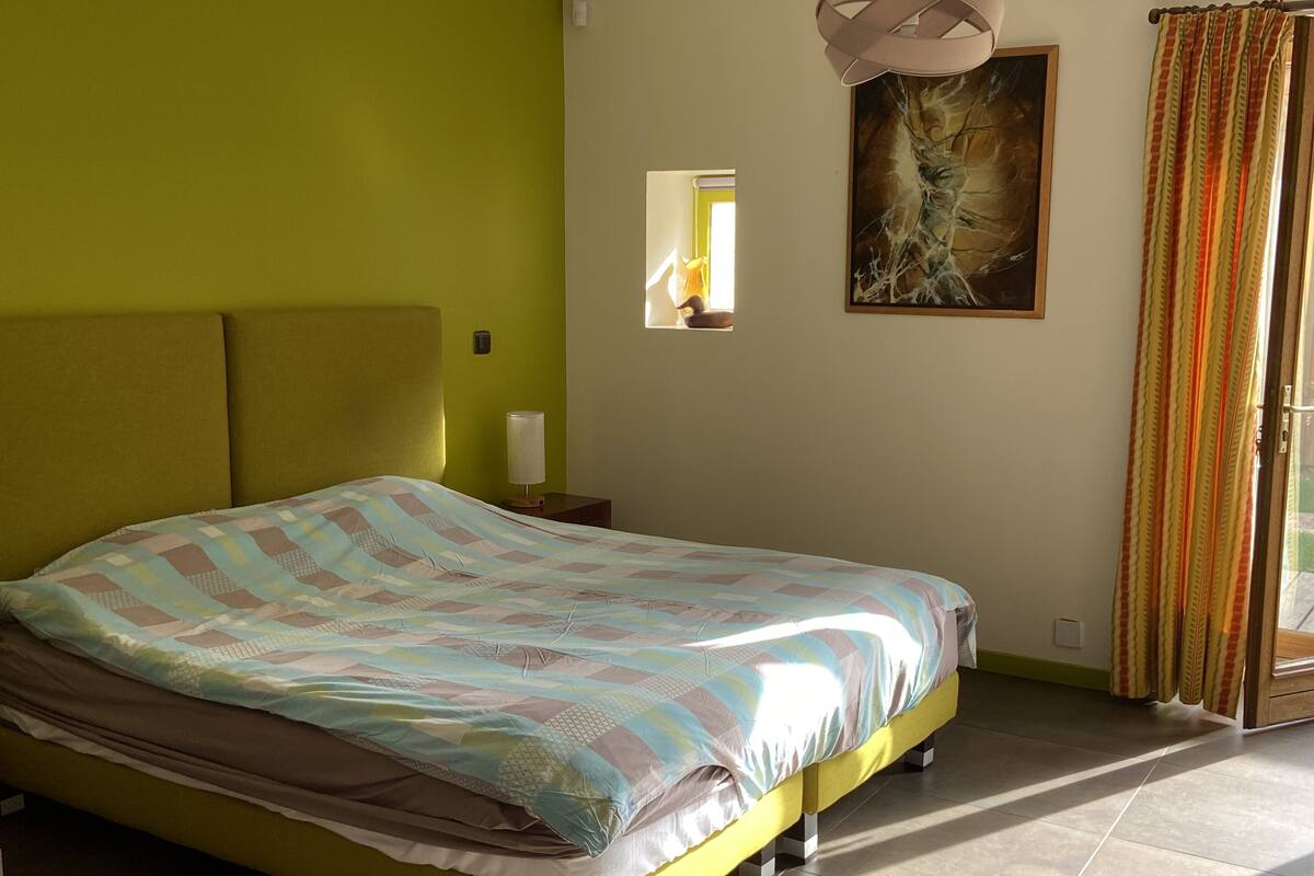A bedroom with a bed, a nightstand, a lamp, a painting, and a window with curtains.