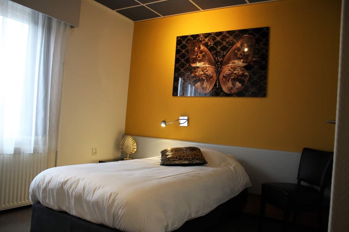 A hotel room with a bed, a chair, a wall-mounted lamp, and a painting.