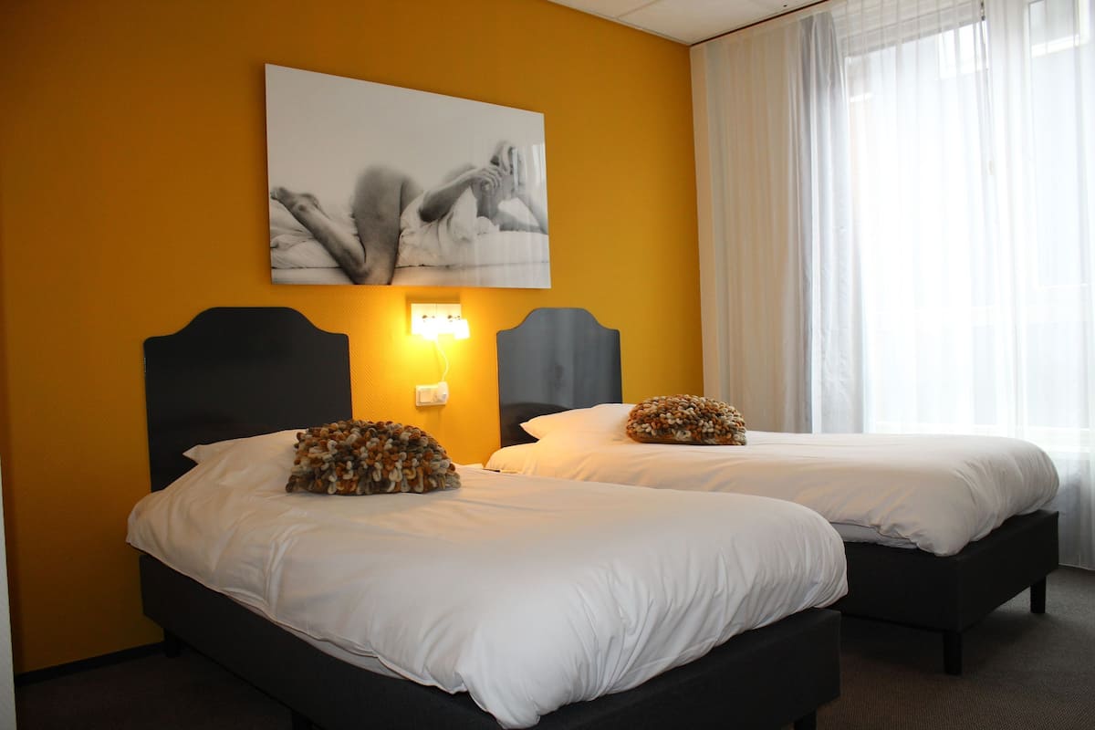 A hotel room with two beds, a large painting on the wall, and a window with sheer curtains.