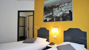 Een hotelkamer met twee bedden, een nachtkastje met een telefoon, een wandlamp en een groot stadsgezicht op een foto.