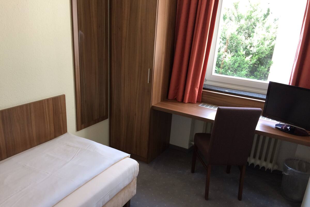 Une chambre à coucher avec un lit, un bureau avec un ordinateur, une chaise et une fenêtre avec des rideaux.