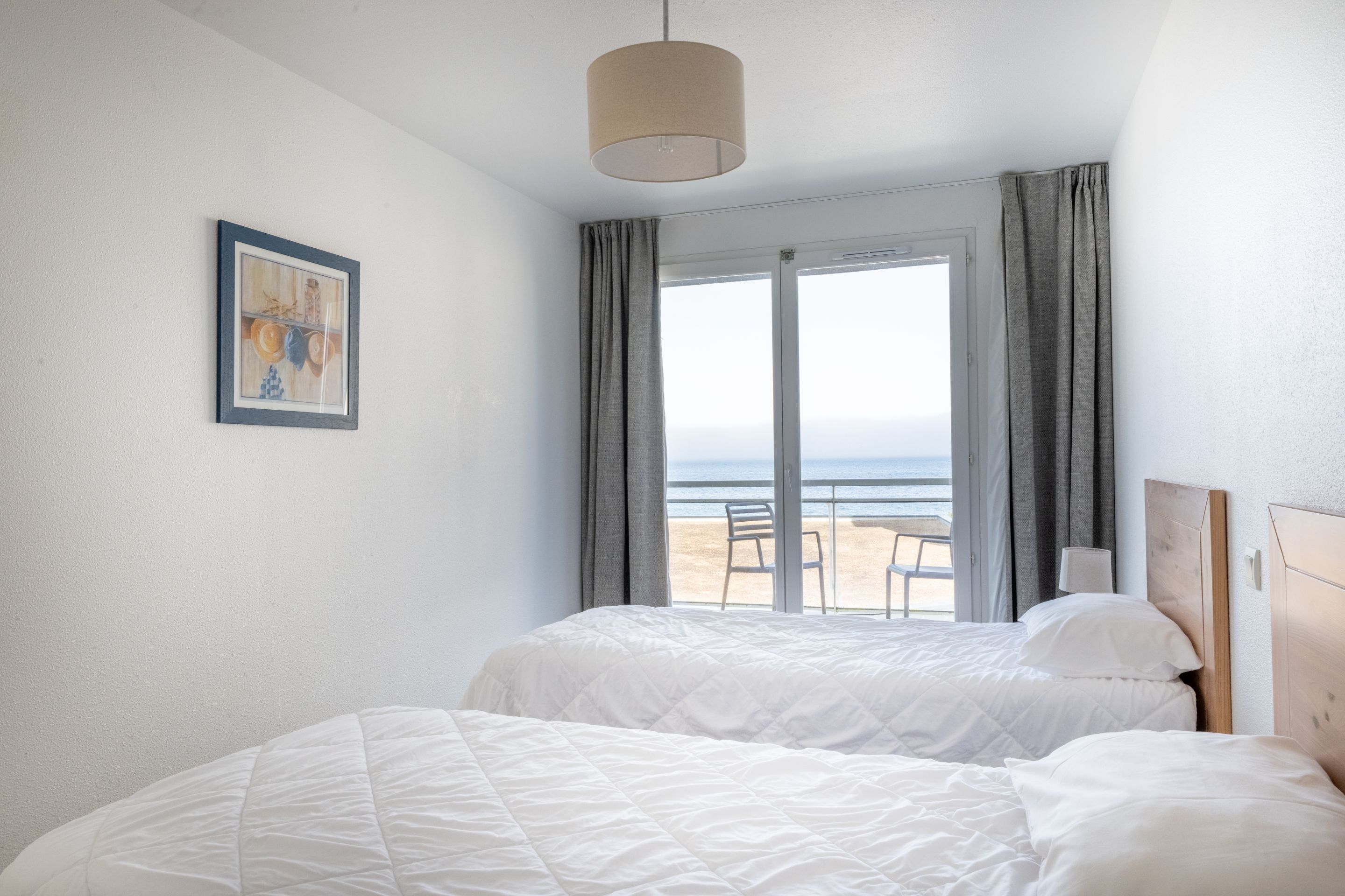 Habitación de hotel con dos camas, un ventanal con vistas a la playa, un cuadro en la pared y una lámpara de techo.