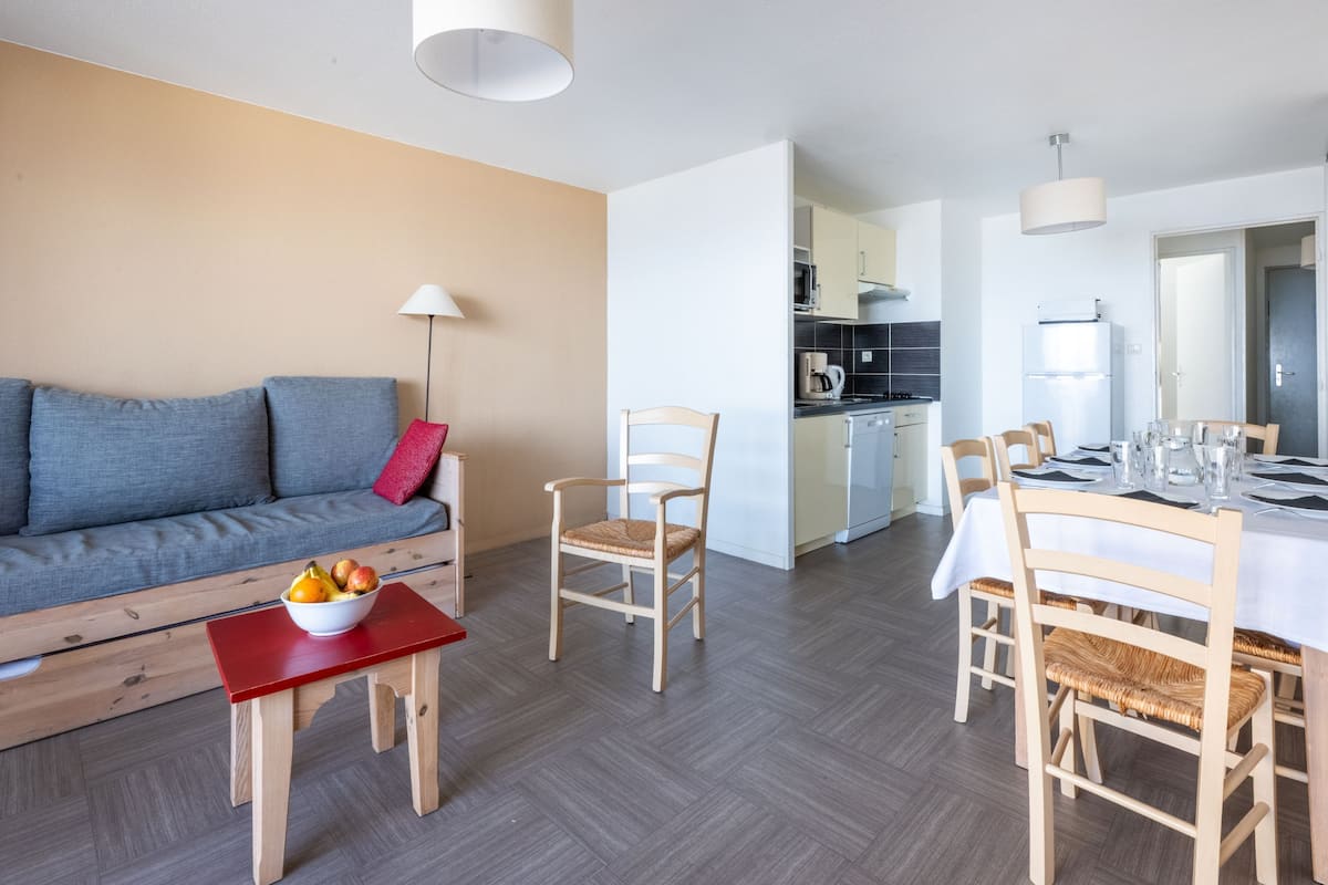 Una sala moderna con un sofá, una mesa de comedor, sillas, una pequeña mesa con fruta y una cocina al fondo.
