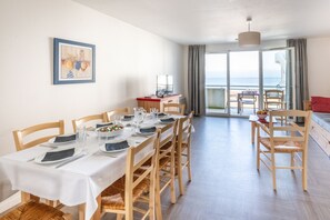 A dining area with a table set for four, wooden chairs, a television on a stand, and a view of the sea through large windows. - Résidence Odalys Le Domaine des Roches Jaunes (Plougasnou)