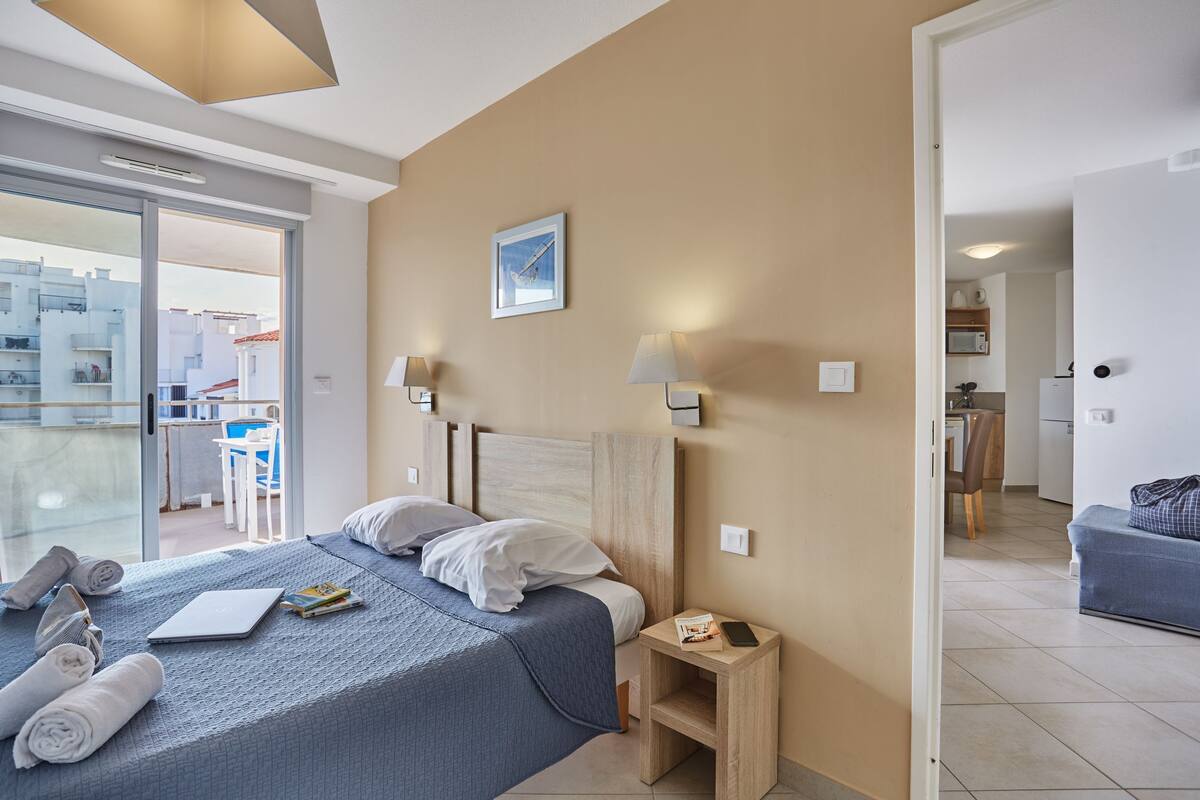 Ein Hotelzimmer mit Bett, Nachttischlampen, einem kleinen Tisch, Blick durch eine Glastür und einem sichtbaren Küchenbereich durch eine offene Tür.