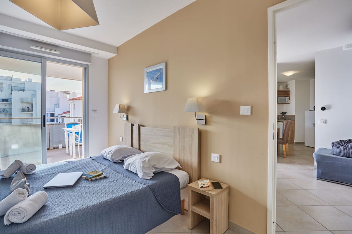 A hotel room with a bed, bedside lamps, a small table, a view through a glass door, and a kitchen area visible through an open door.