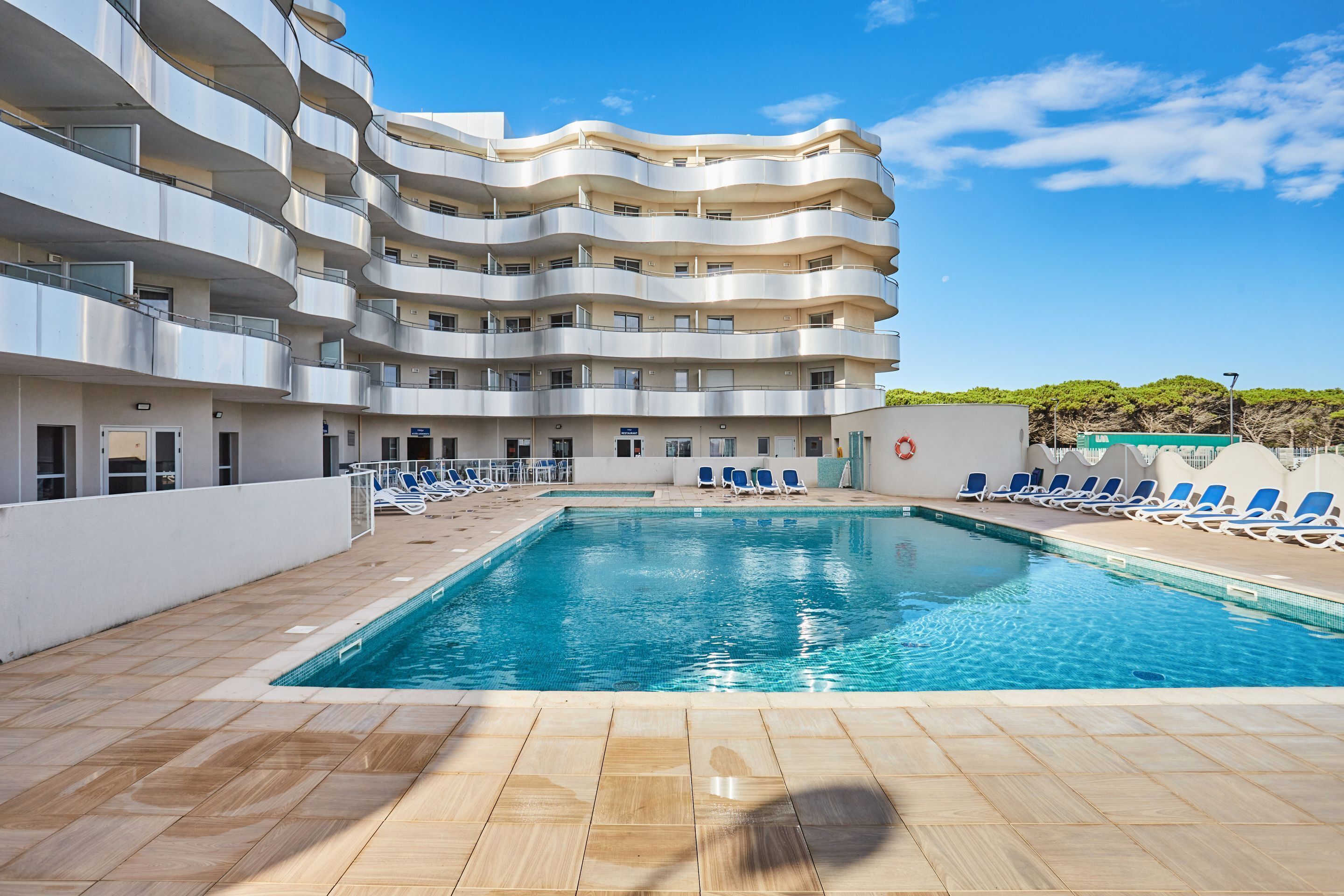 A modern hotel with a swimming pool, multiple balconies, and a clear sky.