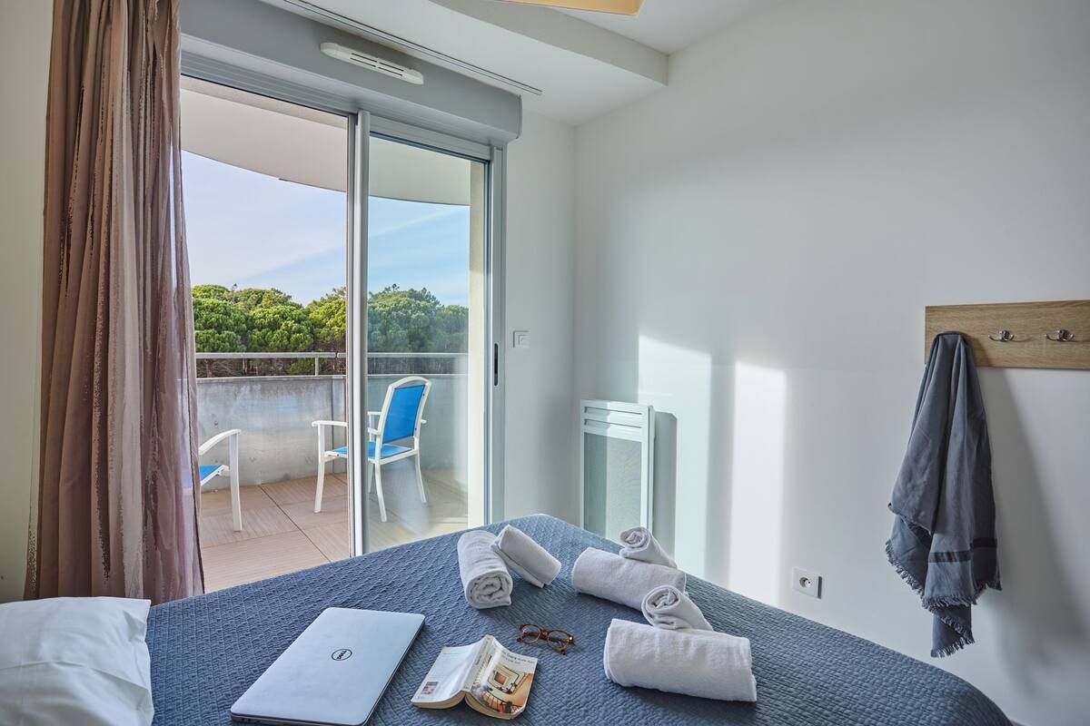 Ein Hotelzimmer mit einem Bett, einem Laptop, einem Buch und einer Brille auf dem Bett. Ein Balkon mit einem Stuhl und Blick ins Grüne ist vorhanden.