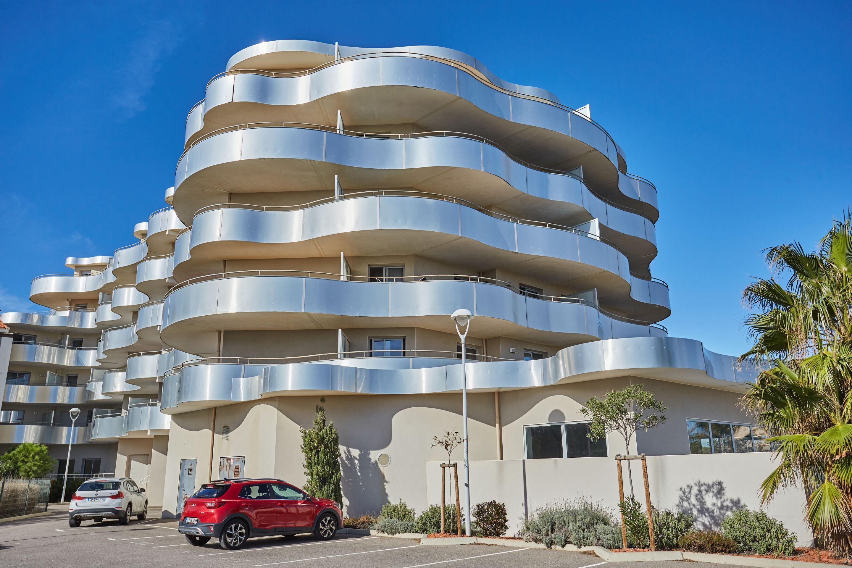 Un immeuble moderne à plusieurs étages, doté de balcons, d’un parking et surplombant un ciel bleu azur.