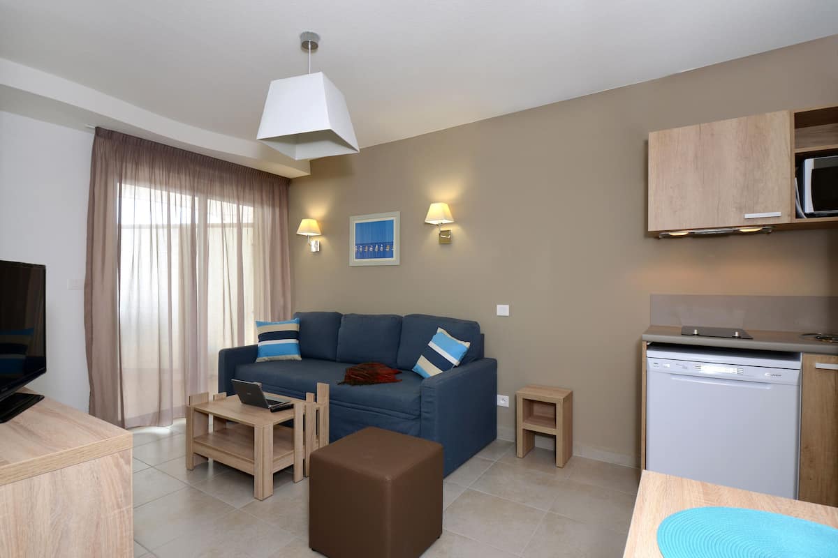 Ein modernes Wohnzimmer mit Sofa, Couchtisch und Fernseher. Es gibt eine Küchenzeile mit Mikrowelle und Geschirrspüler.