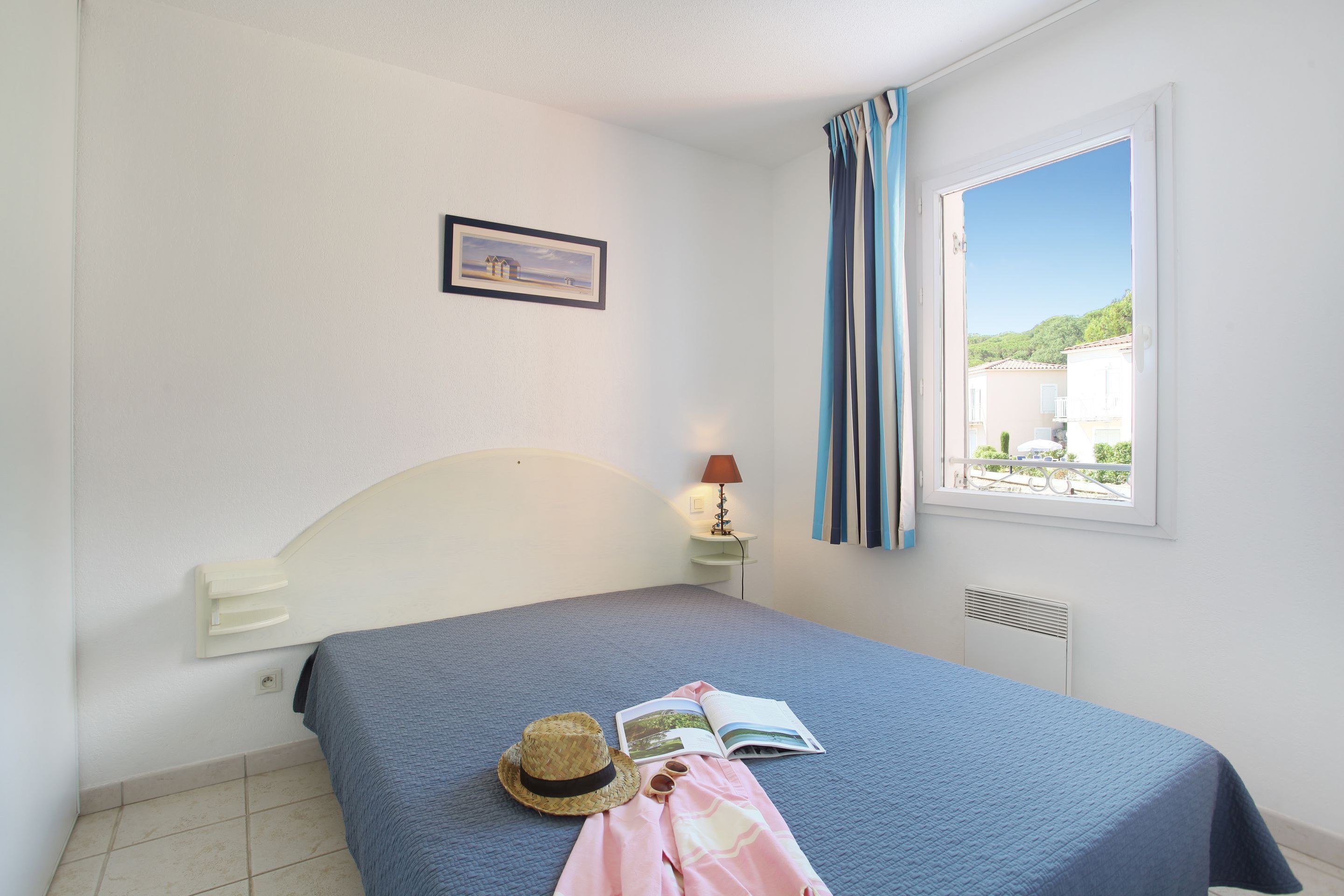 A bedroom with a bed, a nightstand, a lamp, a window with curtains, and a framed picture on the wall.