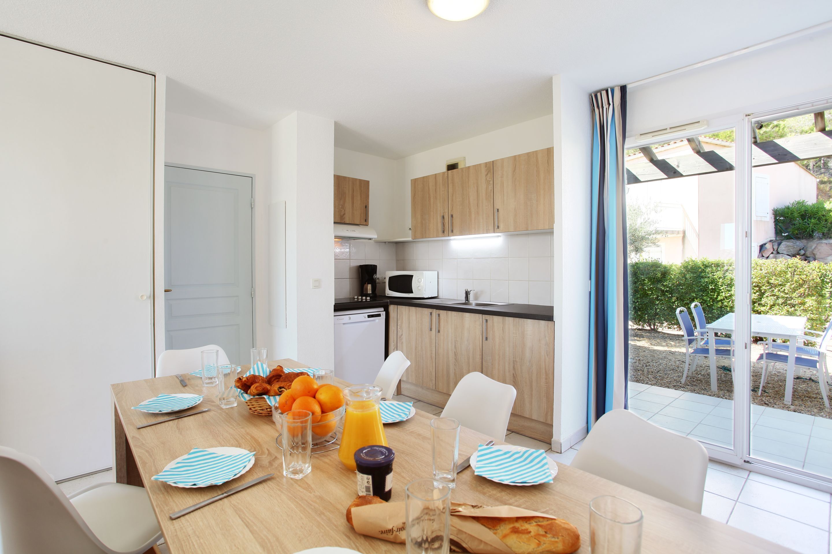 A modern kitchen with wooden cabinets, a microwave, and a dishwasher. There is a dining table set with plates, glasses, and a basket of oranges.