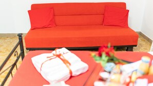 A red sofa bed with matching pillows, a small table with a towel and toiletries, and a flower arrangement. - Terrazze Italiane City Center (Verona)