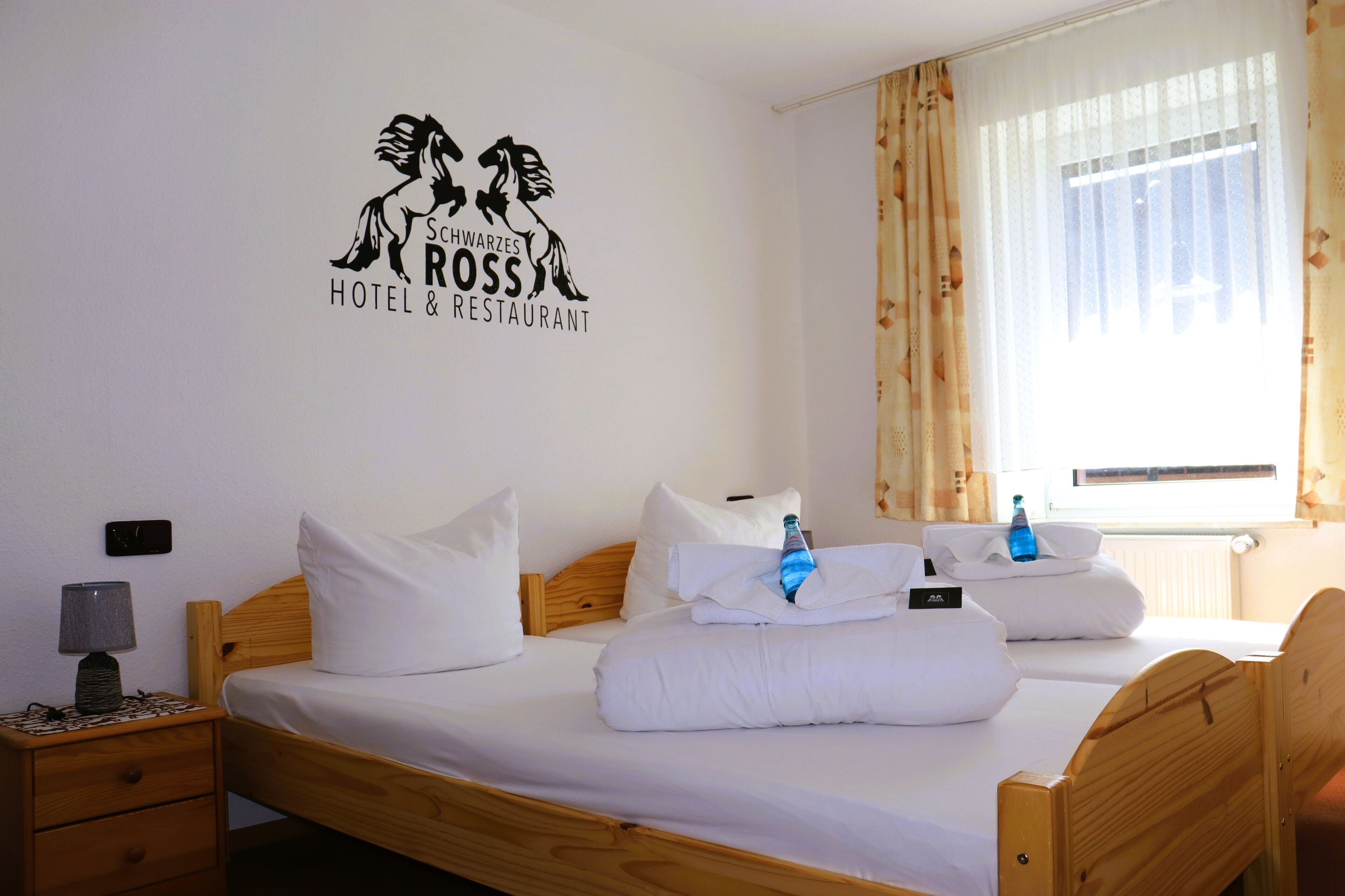 Ein Hotelzimmer mit einem hölzernen Bett, weißer Bettwäsche, einer Wanddekoration, einem Nachttisch mit Lampe und einem Fenster mit Vorhängen.