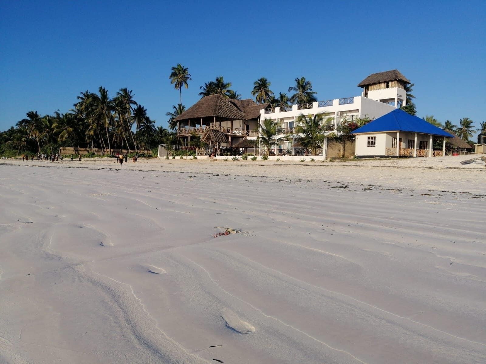 A beach with a hotel featuring a thatched-roof structure, a blue-roofed building, and palm trees.